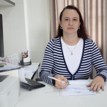 Foto de  Josilene Wosniak de Andrade, coordenadora da Psicologia. Ela est sentada  mesa, de frente para a foto, com um leve sorriso. Com a mo direita, ela segura uma caneta como se estivesse escrevendo num papel. A mo esquerda segura um papel delicadamente. Ela tem cabelos avermelhados, compridos.