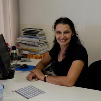 a foto mostra a diretora do CAEE e da Escola Osny Macedo Saldanha sentada  sua mesa,  direita da foto, olhando para foto e sorrindo. Ela est com os dois braos sobre a mesa, tendo a mo esquerda sobre a direita. Tem cabelos pretos e longos, semi-presos.