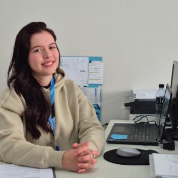 Foto de Luana do Vale que  auxiliar administrativa e responsvel pelo quadro associativo do IPC. Luana tem pele clara, cabelos longos e escuros, levemente ondulados. Ela usa um moletom bege claro e um crach azul pendurado no pescoo. Atrs dela h uma parede branca, um calendrio de papel fixado e alguns papis organizados. Sobre a mesa, h um notebook aberto com um monitor acoplado, uma garrafa de gua, um mouse sobre um mousepad, papis organizados e uma impressora ao fundo.