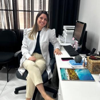 Foto de Keyla Palestino, respons�vel pelo setor de fisioterapia do IPC. Keyla tem pele clara, cabelos lisos e castanhos, com mechas mais claras nas pontas. Ela est� sentada em uma cadeira de escrit�rio, sorrindo, e veste um jaleco branco sobre uma blusa preta e cal�a bege clara. Usa sapatos marrons e est� em uma sala bem iluminada, com cortinas escuras ao fundo. Sobre a mesa ao lado, h� um computador, cadernos, uma bola de fisioterapia, uma cesta com materiais e uma capa de celular rosa.