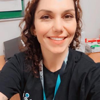 Foto de Padwa Cristina, coordenadora do setor de enfermagem do IPC. Padwa tem pele clara, cabelos castanhos escuros e cacheados, usa uma tiara branca e est� sorrindo. Ela veste uma camiseta preta com o logotipo do IPC e um crach� azul pendurado no pesco�o. A foto � tirada em um ambiente de trabalho, com uma mesa de madeira ao fundo, pap�is organizados, uma parede clara e parte de um quadro verde vis�vel.