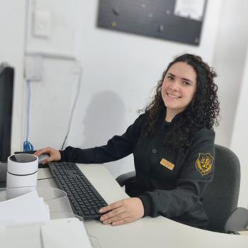 Foto de Kauane Lopes, nutricionista do IPC. Kauane tem pele clara, cabelos cacheados e castanhos escuros, e est� sorrindo. Ela veste um jaleco preto com o bras�o dourado do IPC na manga e uma plaquinha de identifica��o no peito. Est� sentada em uma mesa de trabalho, com uma das m�os sobre o teclado de um computador e pap�is organizados � frente. Ao fundo, h� uma parede branca e um quadro escuro fixado na parede.