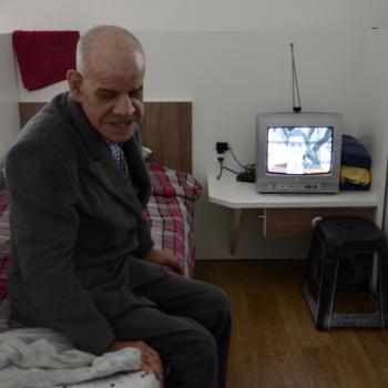 a imagem mostra um morador dentro do seu quarto. Ele est sentado aos ps da cama, sobre um cobertor xadrez de vermelho e marrom, com as pernas para a lateral da cama. Est com a cabea voltada para a esquerda, no sentido da foto. Ele sorri. Ao lado da cabeceira da cama, h um criado-mudo com uma pequena televiso, que est ligada. O quarto  fechado com paredes baixas e brancas.