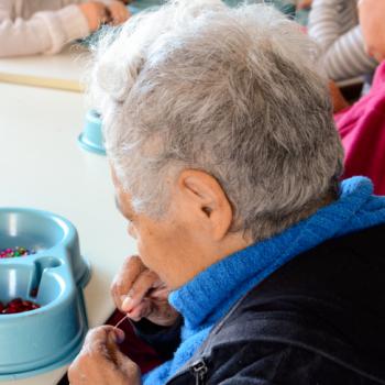 a imagem mostra uma moradora idosa de costas, sentada  mesa branca, participando de uma atividade de artesanato com miangas. Ela est colocando uma bolinha vermelha na linha. Tem  sua rente um recipiente azul repleto de miangas coloridas. Ao lado direito dela esto outros moradores, que aparecem em segundo plano, desfocados, sem mostrar o rosto.