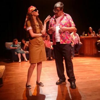 a imagem mostra uma cena da pea teatral, com uma atriz e um ator conversando. A atriz est falando e o ator, ouvindo. Ela tem o brao esquerdo apoiado na horizontal, passando por cima da barriga, de um lado a outro, e o cotovelo direito apoiado nele, na vertical. Ela usa culos. O ator est segurando a camisa colorida que veste, que est aberta e tem por baixo dela uma camiseta branca. Ele est srio ouvindo a atriz. Ao fundo da cena, h atores sentados num sof listrado e em cadeiras.
