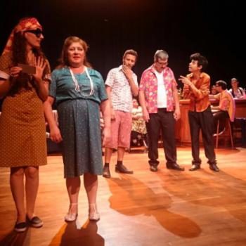 a imagem mostra uma cena da pea teatral, com todos os atores em p, em diferentes cenas menores dentro da cena principal. A principal traz duas atrizes conversando. A da direita segura um papel pequeno com a mo esquerda, gesticula com a mo direita e fala com a atriz que est ao seu lado esquerdo (direito da foto). Esta, por sua vez, sorri e escuta o que est sento dito. Atrs delas, dois atores conversam e um as observa. Atrs deles, outros atores esto sentados a uma mesa de bar, alguns esto em p atrs da mesa. Eles conversam.