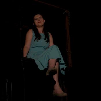 a imagem mostra uma cena da peça teatral, com uma atriz sentada com as pernas cruzadas, sorrindo, braços ao lado do corpo e cabeça ligeiramente tombada para a direita. A imagem tem ângulo de baixo para cima.