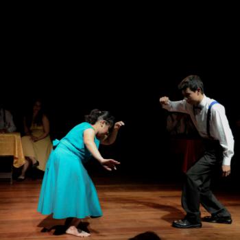 a imagem mostra uma cena da pea teatral, com uma atriz e um ator em p, danando. Ambos olham para baixo. Ao fundo,  esquerda da foto, h trs atores sentados a uma mesa, observando a cena principal.