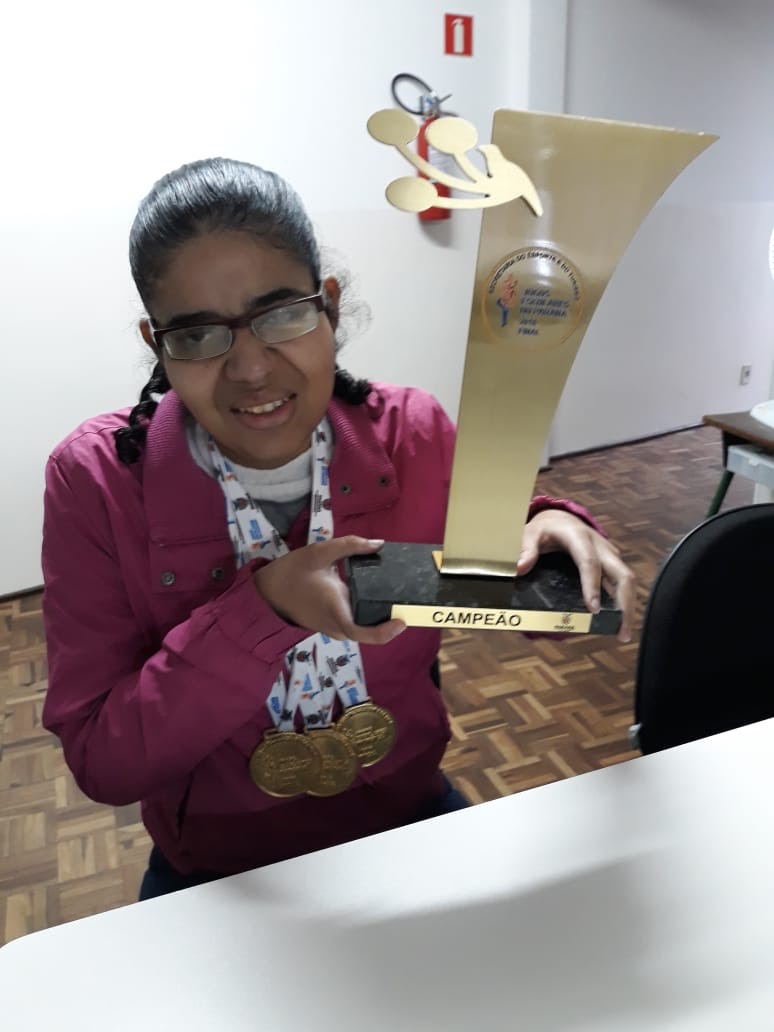 A foto mostra Dayane sentada a uma mesa branca, tendo três medalhas de ouro penduradas no pescoço e em segurando com as duas mãos, do lado esquerdo do corpo (direito da foto) o troféu dourado. Ela sorri enquanto posa para a foto.